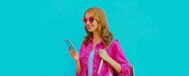 Akıllı telefonlu, pembe ceketli, mavi arka planda güneş gözlüklü şık bir genç kadının portresi.