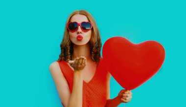 Güzel bir kadının portresi, kırmızı kalp şeklinde balonlar uçuşan dudaklar, tatlı bir hava öpücüğü gönderen mavi bir arka planda güneş gözlüğü takan bir kadın.