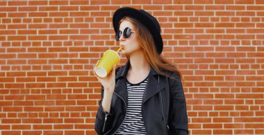 Tarz sahibi genç bir kadının portresi. Meyve suyu içiyor. Tuğla duvarın arkasında siyah bir kaya tarzı giyiyor.