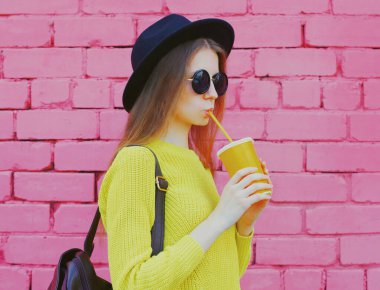 Şık bir genç kadının portresi. Meyve suyu içiyor. Pembe tuğladan bir arka planda sarı örülü süveter giyiyor.