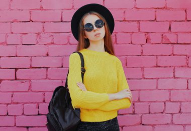 Pembe tuğladan bir arka planda sarı örülü süveter giyen zarif genç bir kadının portresi.