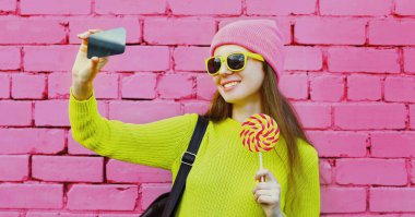 Pembe tuğla duvarda lolipopla telefonla selfie çeken genç bir kadının portresi.
