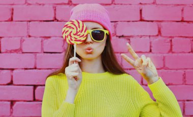 Renkli lolipopu olan sarı süveter giyen, pembe arka planda şapkası olan bir kızın portresi.