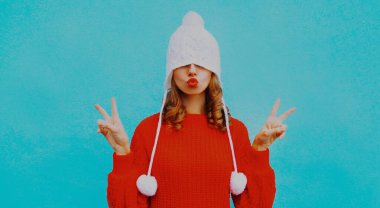 Genç bir kadının portresi kırmızı dudaklar üflüyor, hava öpücüğü gönderiyor, beyaz şapkalı, ponponlu, mavi arka planda kırmızı örülü süveter giyiyor.