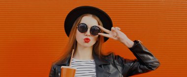 Meyve suyu bardağında, siyah yuvarlak şapkalı, turuncu arka planda ceketli şık bir genç kadının portresi.