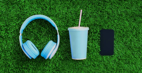 Close up blue wireless headphones, smartphone, cup of juice on a green grass background, top view