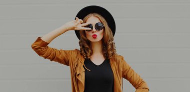 Portrait of beautiful young woman blowing red lips wearing a black round hat, brown jacket on a gray background