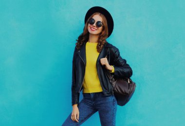 Mavi arka planda siyah bir şapka ve sırt çantası takan gülümseyen genç bir kadının portresi.