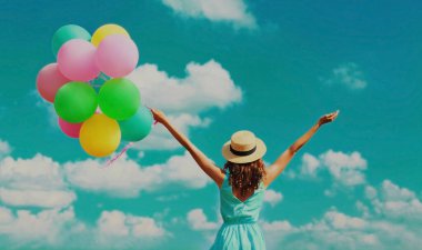 Mutlu gülümseyen genç kadın, mavi gökyüzü bulutları üzerinde yaz hasır şapkası takan renkli balonlarla dolu.