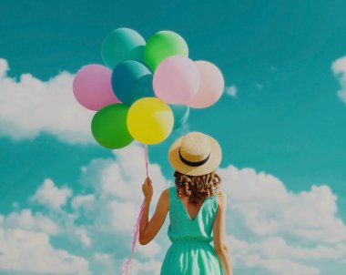 Arka planda mavi gökyüzü arka planında yaz hasır şapkası takan renkli balonları olan genç bir kadın var.
