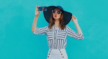 Güzel genç bir kadının portresi akıllı telefondan fotoğraf çekiyor. Siyah yuvarlak bir şapkası var. Mavi arka planda beyaz çizgili bir gömlek.