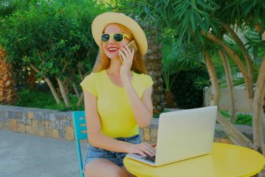 Yaz tatilinde masada otururken dizüstü bilgisayarla telefonla arayan mutlu genç bir kadın.