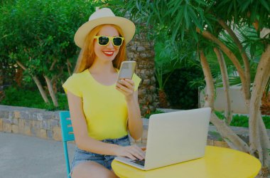 Yaz tatilinde masada otururken telefonuyla ve laptopuyla gülümseyen genç bir kadın.