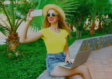 Bilgisayarlı mutlu genç bir kadın, bir yaz parkında cep telefonuyla fotoğraf çekiyor.