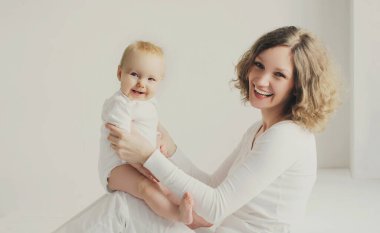 Mutlu gülümseyen genç anne, küçük bebeğiyle birlikte beyaz odada oynuyor.