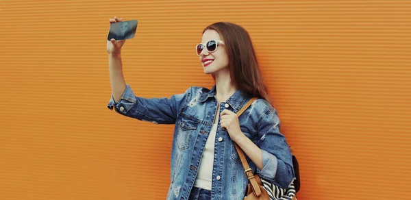 用智能手机在橙色背景下穿着斜纹棉布夹克拍自拍照的女人的画像 — 图库照片