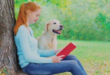 Parktaki bir ağacın yanında Golden Retriever köpeğiyle kitap okuyan genç bir kadın.