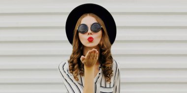 Kırmızı dudaklarını sümküren, siyah yuvarlak şapkalı, beyaz arka planda gömlek giyen genç bir kadının portresi.