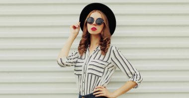 Beyaz arka planda çizgili gömlek ve siyah yuvarlak şapka giyen şık bir genç kadının portresi.
