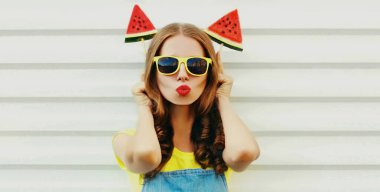 Kırmızı dudaklarını dondurma şeklinde bir karpuz dilimiyle beyaz arka planda sallayan genç bir kadının portresi.