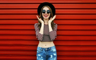 Kırmızı arka planda siyah yuvarlak şapkalı ve güneş gözlüklü mutlu genç kadının portresi.