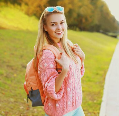 Parkta sırt çantası takan mutlu, genç, sarışın bir kadının portresi.