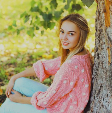 Güz parkında pembe kazak giyen güzel, gülümseyen genç bir kadının portresi.