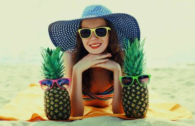 Mutlu gülümseyen genç bir kadının portresi kumsalda uzanmış komik bir ananasla hasır şapka takıyor.