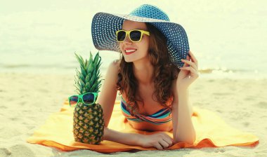 Mutlu gülümseyen genç bir kadının portresi kumsalda uzanmış komik bir ananasla hasır şapka takıyor.