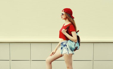 Kırmızı beysbol şapkası ve sırt çantası takan genç bir kadın beyaz arka planda bir şehre bakıyor.