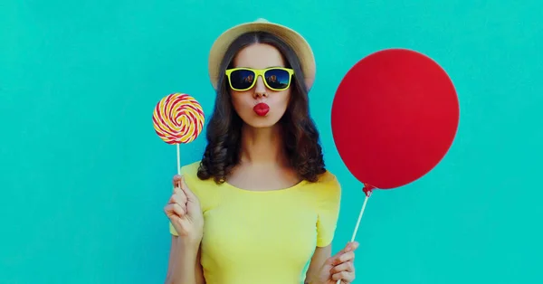 Portrait close up of young woman with lollipop and red balloon blowing a red lips wearing a summer straw hat on a blue background
