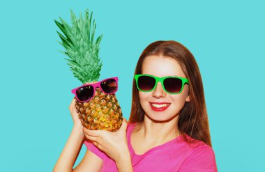 Mavi arka planda izole edilmiş güneş gözlüğü takan ananaslı neşeli genç kadın modelinin portresi.