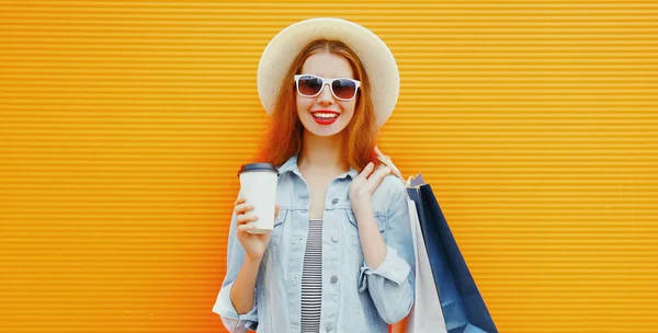 ショッピングバッグやコーヒーカップを身に着けている幸せな若い女性の肖像オレンジの背景に夏のわら帽子 — ストック写真