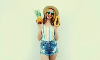 Ananas ve papaya takmış, beyaz arka planda güneş gözlüğü takan mutlu genç bir kadının yaz portresi.