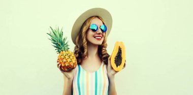 Ananas ve papaya takmış, beyaz arka planda güneş gözlüğü takan mutlu genç bir kadının yaz portresi.