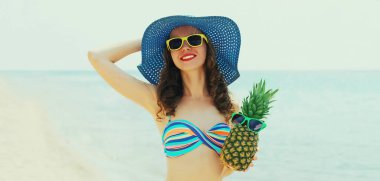 Kumsalda, komik ananaslı, hasır şapkalı, gülümseyen genç bir kadın.