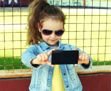 Küçük bir kızın portresi şehrin caddesinde akıllı telefonla selfie çekiyor.
