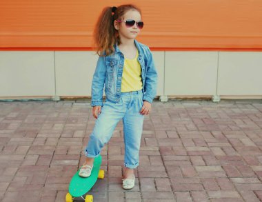 Küçük kız çocuğunun portresi turuncu bir arka planda kaykayla şehirde poz veriyor.