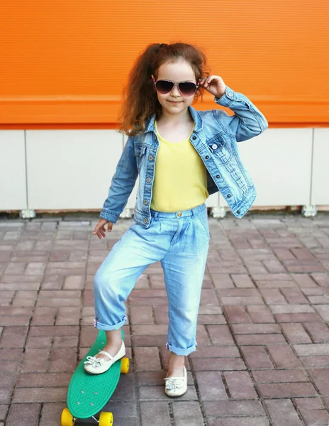 Küçük kız çocuğunun portresi turuncu bir arka planda kaykayla şehirde poz veriyor.