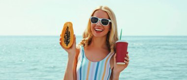 Mutlu gülümseyen genç bir kadının yaz portresi. Elinde papaya ve bir bardak meyve suyu ile bir sahilde.