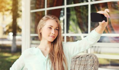 Close up of young blonde woman taking a selfie picture by smartphone sitting at a table in a cafe
