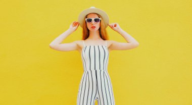 Beyaz çizgili tulum giymiş, sarı bir arka planda poz veren yaz şapkalı şık model bir kadın.