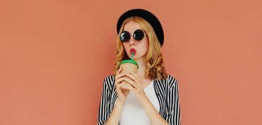 Kahve ya da meyve suyu içen şık bir genç kadının portresi çizgili beyaz gömlek, şapka ve kahverengi arka planda güneş gözlüğü takıyor.