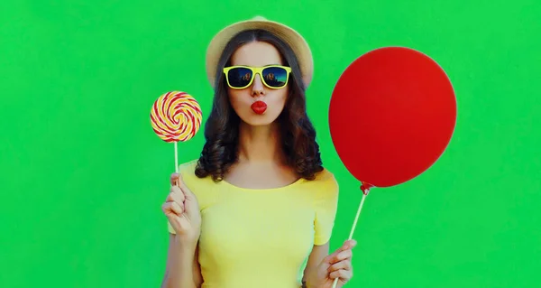 Portrait close up of young woman with lollipop and red balloon blowing her lips wearing a summer straw hat on green background