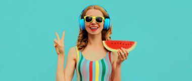 Mavi arka planda sulu bir karpuz dilimiyle müzik dinlerken kulaklıkla poz veren neşeli, mutlu genç kadın modelinin renkli yaz portresi.