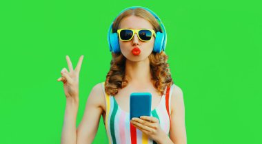 Yeşil arka planda telefonuyla kulaklıkla müzik dinleyen şık genç bir kadının renkli yaz portresi.