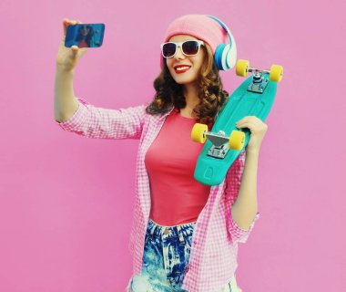 Summer colorful portrait of stylish modern young woman taking a selfie by smartphone with skateboard and headphones on pink background
