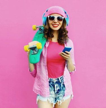 Summer colorful portrait of happy smiling young woman listening to music in headphones with smartphone and skateboard on pink background