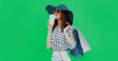 Portrait of beautiful young woman drinking coffee with shopping bags wearing a black round summer hat on green background