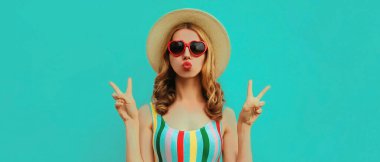 Dudaklarını silen ve yaz hasır şapkası takan, mavi arka planda güneş gözlüğü takan güzel bir kadın modelin portresi.